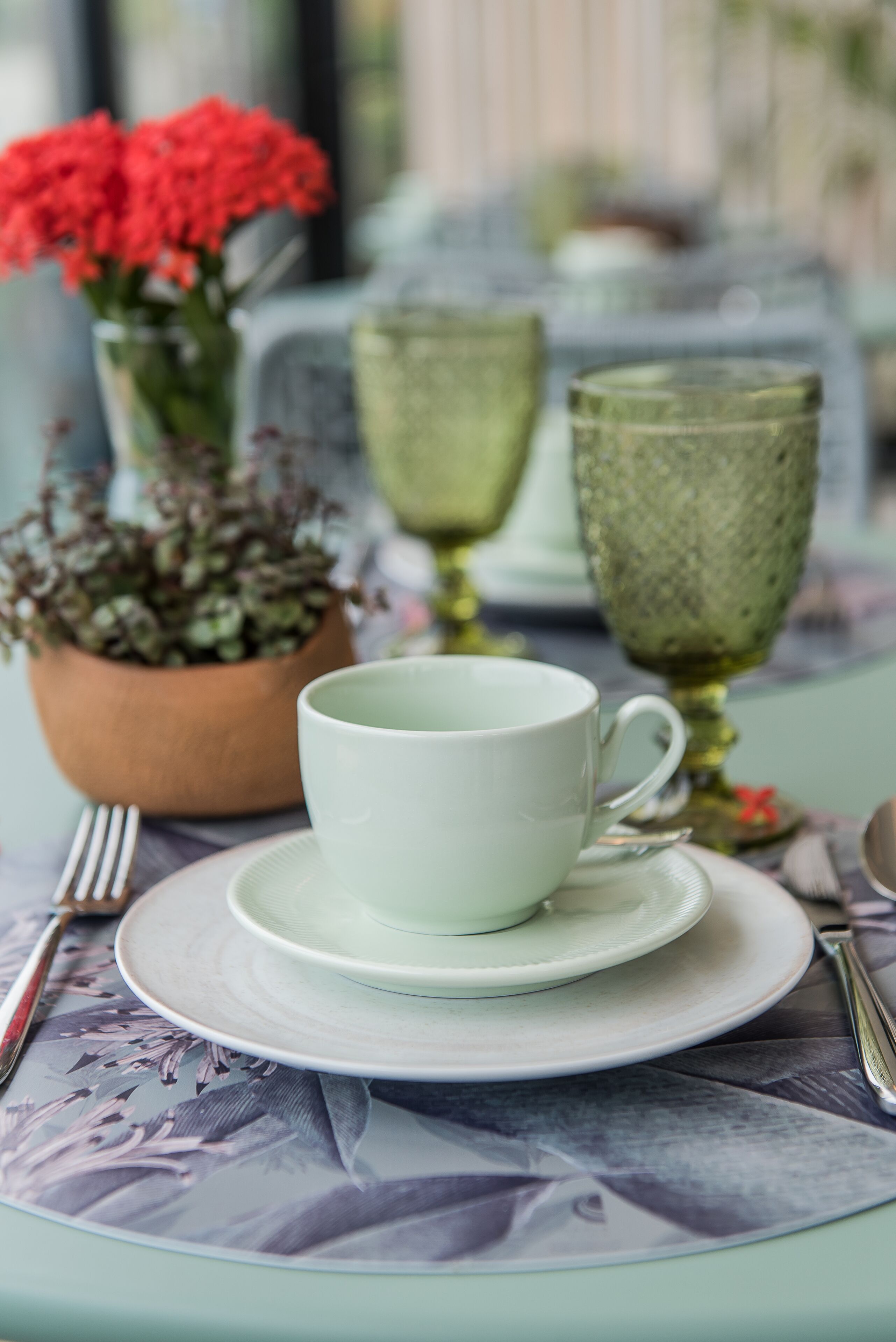 A table set with a white cup and saucer, a green glass, and a potted plant with red flowers.