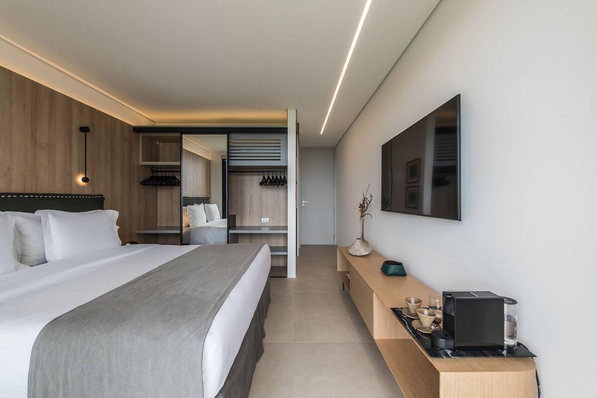 A modern hotel room with a large bed, a bedside table with a coffee maker, a mirror, and a wardrobe with a sliding door.