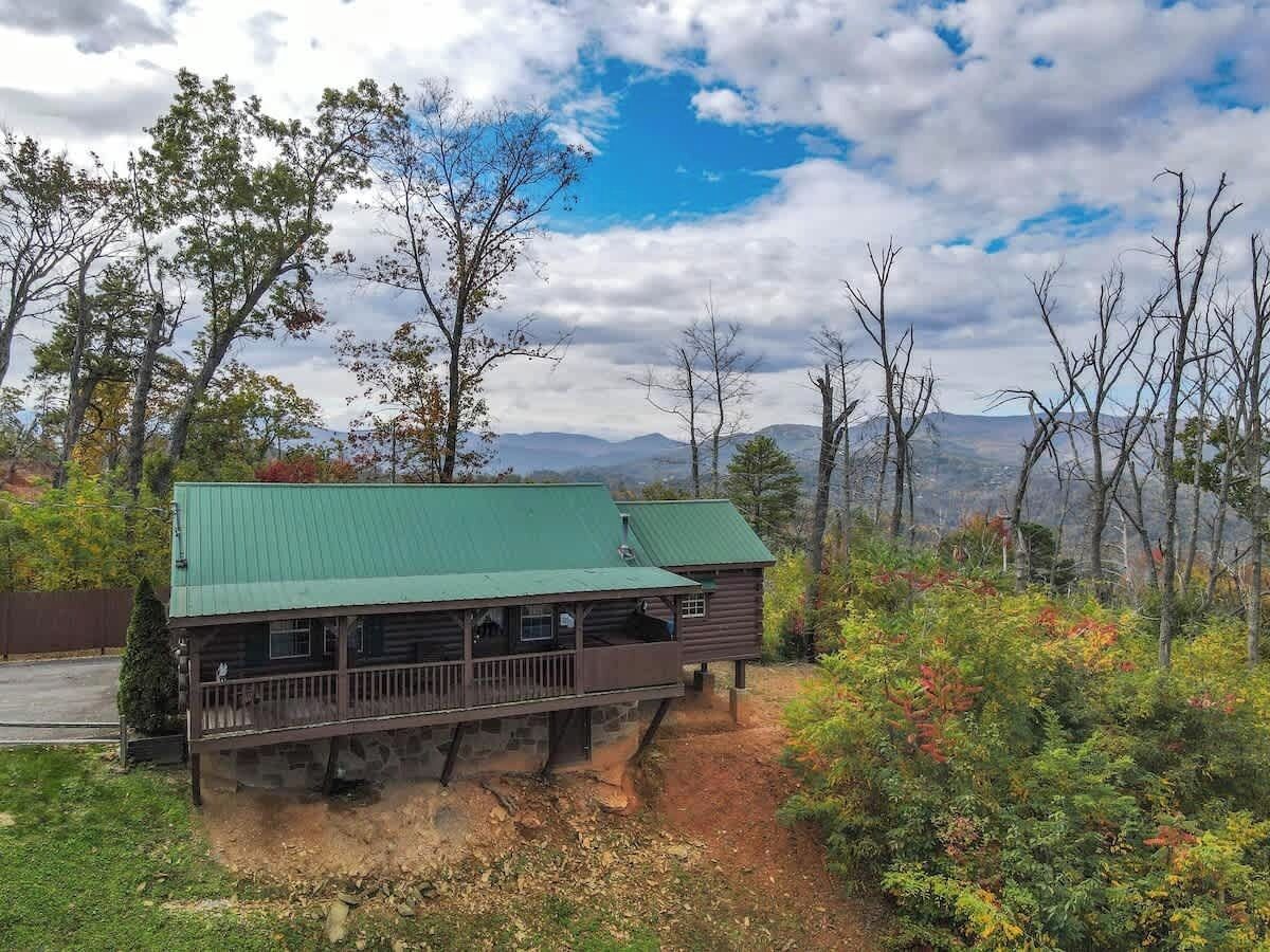 Una baita rustica con tetto verde, un portico e fondamenta in pietra, immersa tra alberi con montagne sullo sfondo.