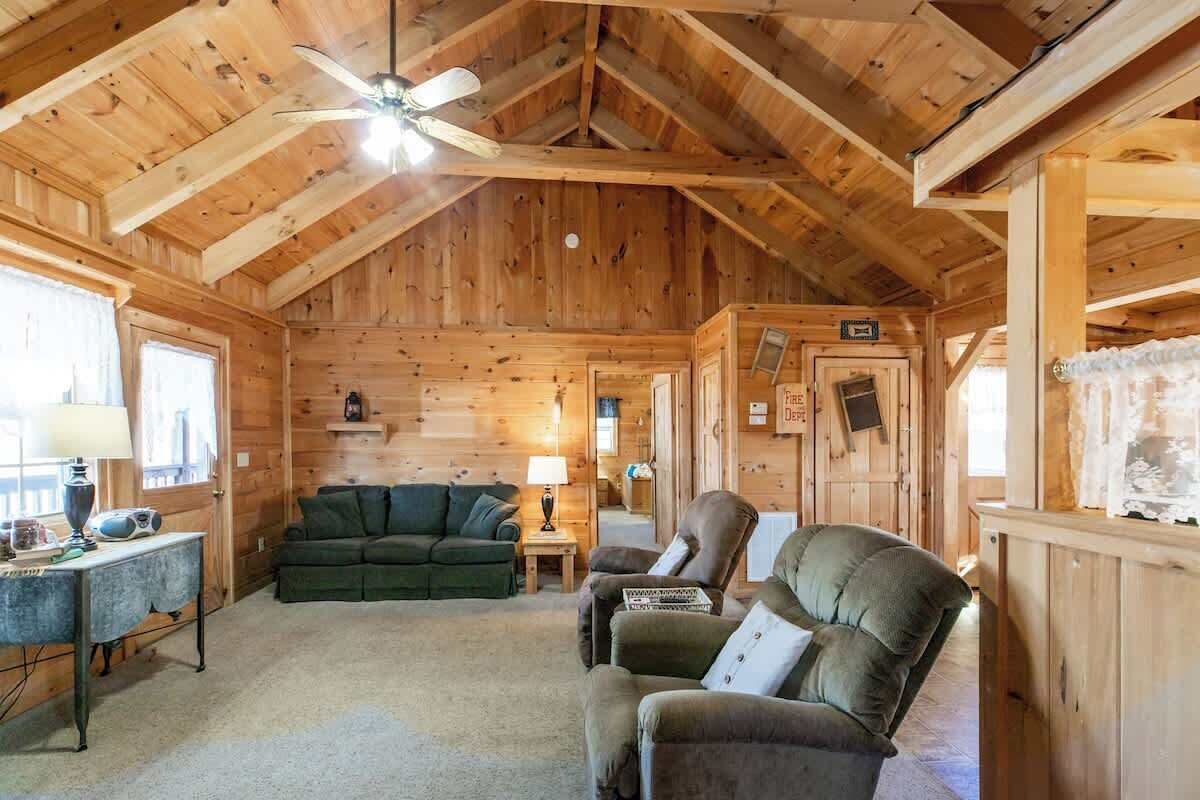 Accogliente interno di una baita in legno con soffitto alto, pareti in legno, un divano, una poltrona e un tavolino con una lampada.
