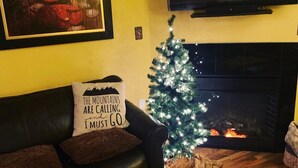 A cozy living room with a fireplace, a black leather couch, a small decorated Christmas tree, and a painting on the wall.
