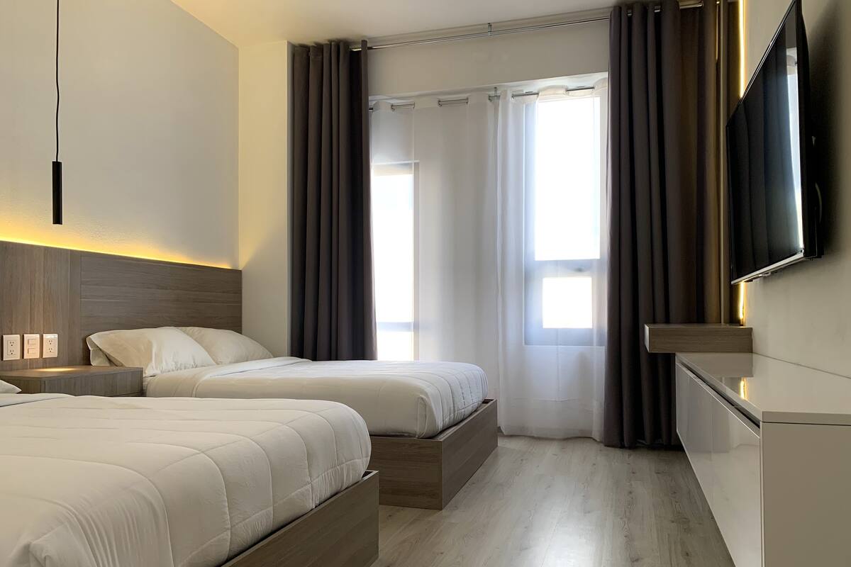 A hotel room with two beds, a TV, a bedside table, and a window with curtains.