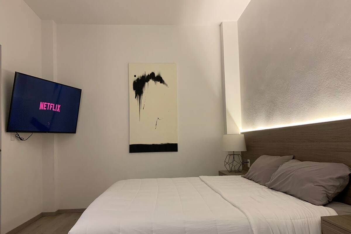 A hotel room with a bed, a TV displaying 'NETFLIX', a wall art piece, and a bedside table with a lamp.