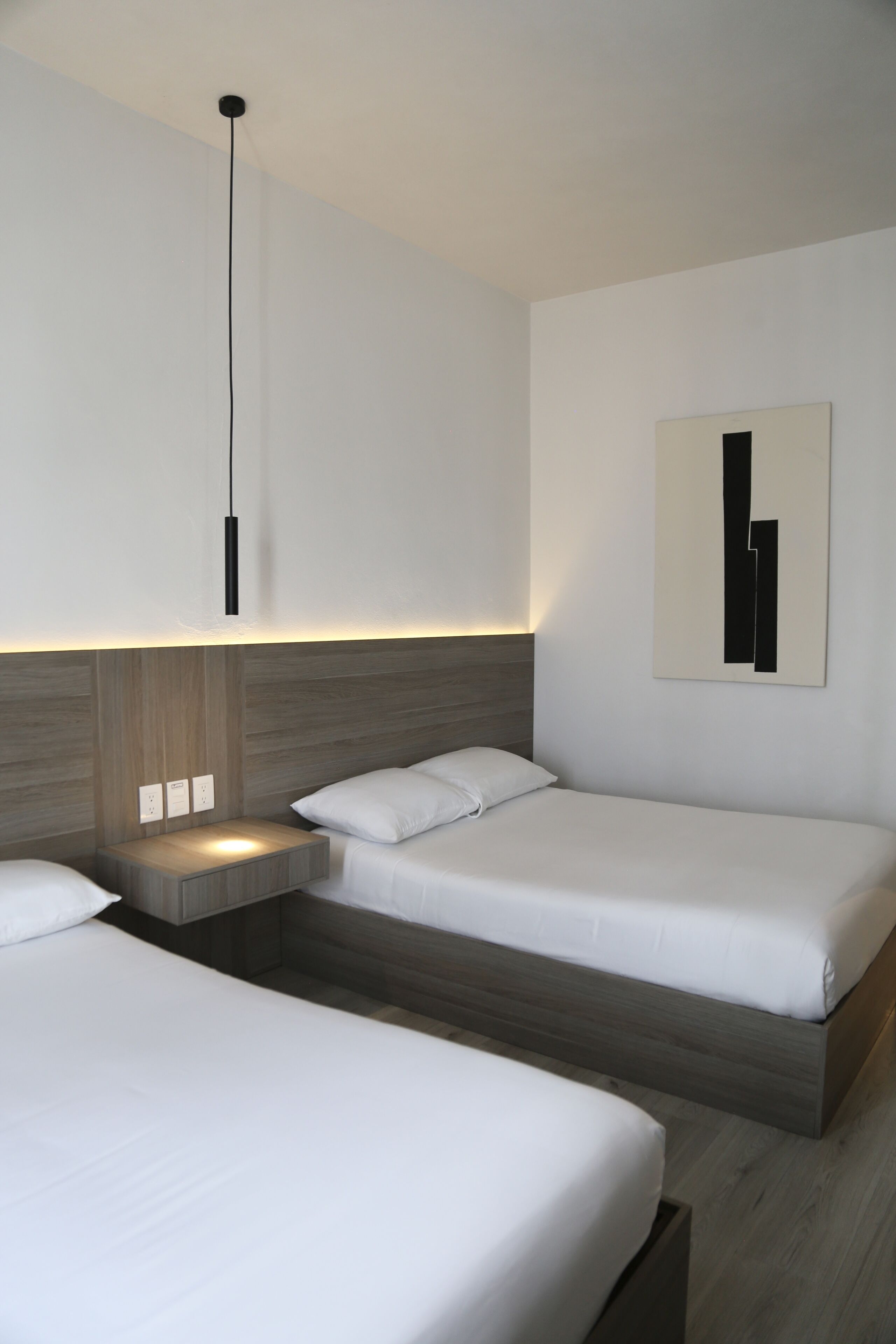 A modern hotel room with two single beds, a bedside table with a lamp, and a wall-mounted artwork.