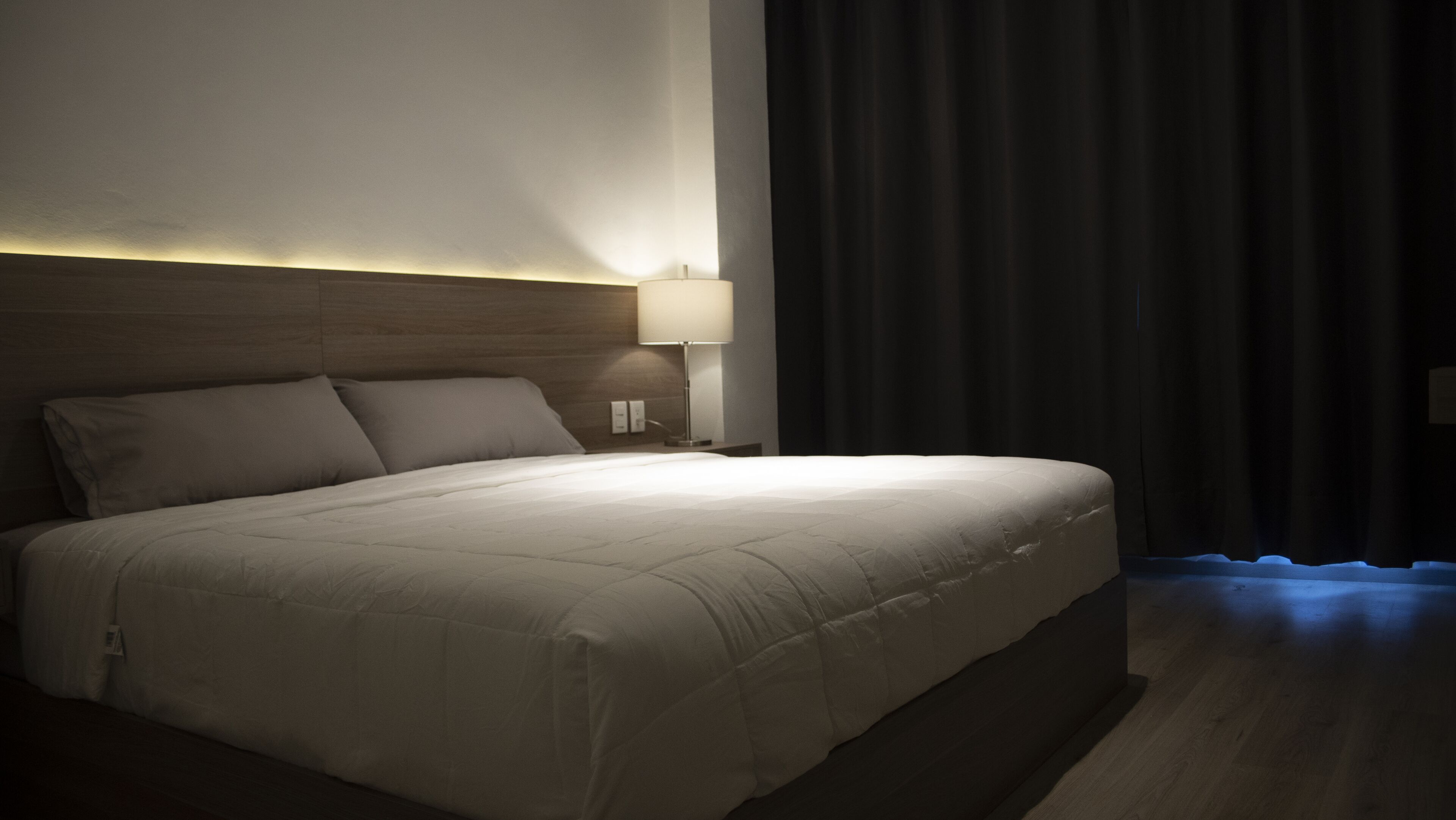 Un lit bien fait, avec une tête de lit, une lampe de chevet et un rideau foncé.