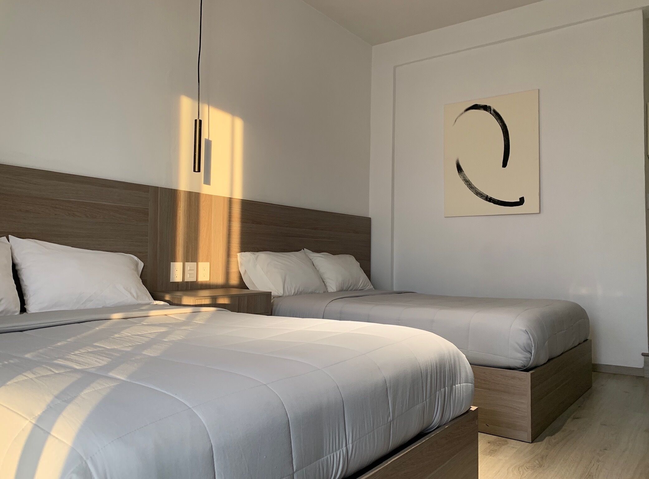 A modern bedroom with two beds, a wooden headboard, a wall-mounted artwork, and a pendant light.