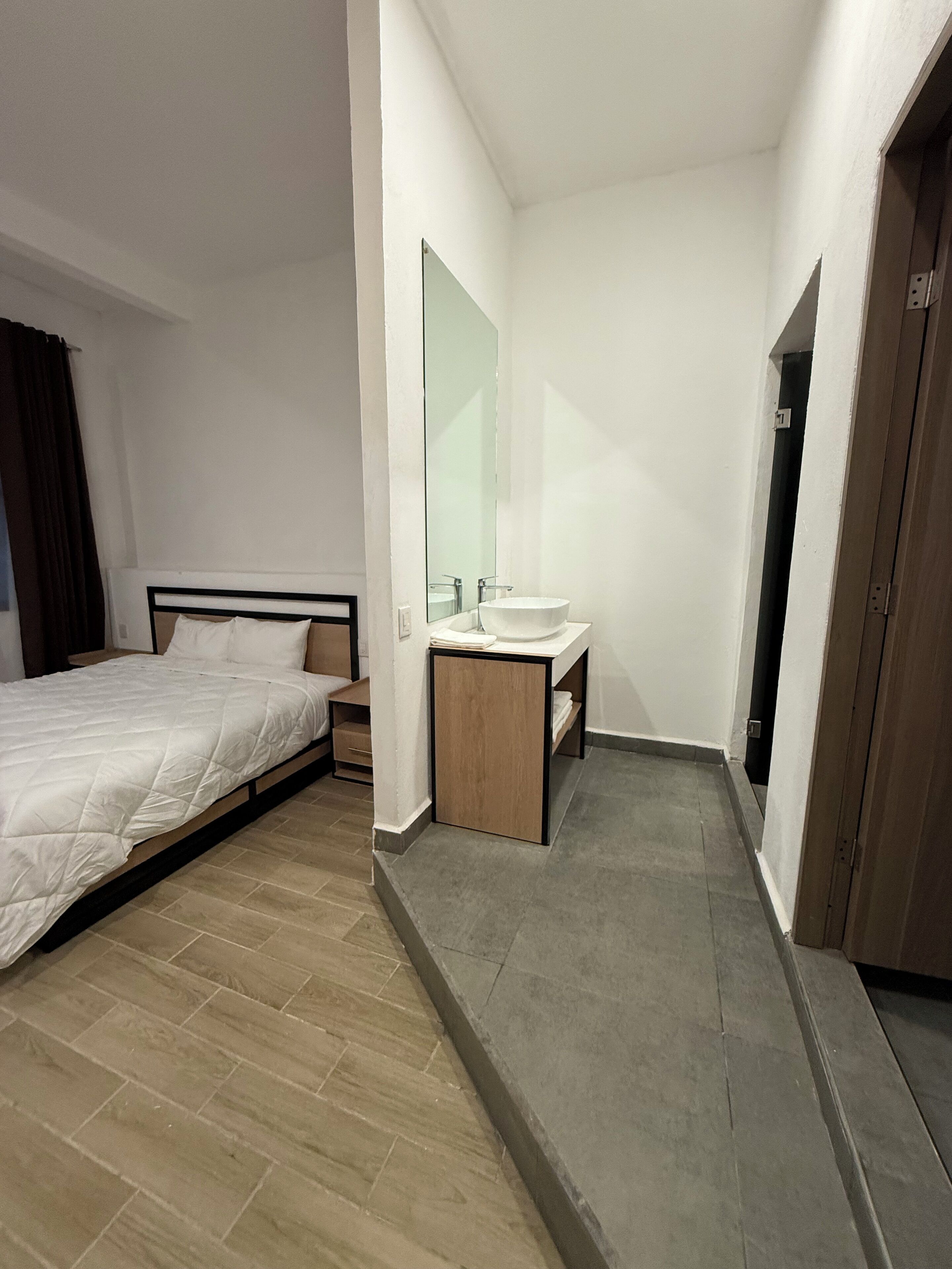 Una habitación de hotel con una cama, una mesita de noche de madera, un lavamanos blanco y un espejo.