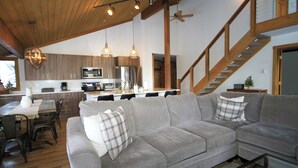A modern living room with a grey sectional sofa, wooden ceiling beams, and a kitchen in the background. - Mountainback #116, Loft,Corner-no pets accepted (Mammoth Lakes)