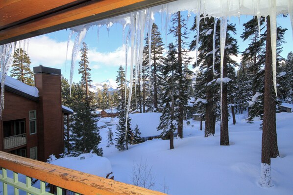 A snowy landscape with icicles hanging from a balcony railing, surrounded by snow-covered trees and a building with a brick facade. - Mountainback #116, Loft,Corner-no pets accepted (Mammoth Lakes)