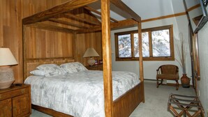 Ein hölzernes Himmelbett mit weißer Bettdecke, ein Nachttisch mit Lampe, ein Sessel und ein Fenster mit Rollläden.