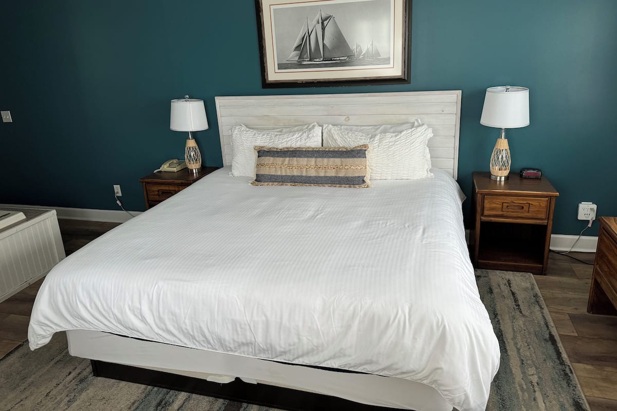 A neatly made bed with white linens, two bedside tables with lamps, a framed picture on the wall, and a radiator under the bed.
