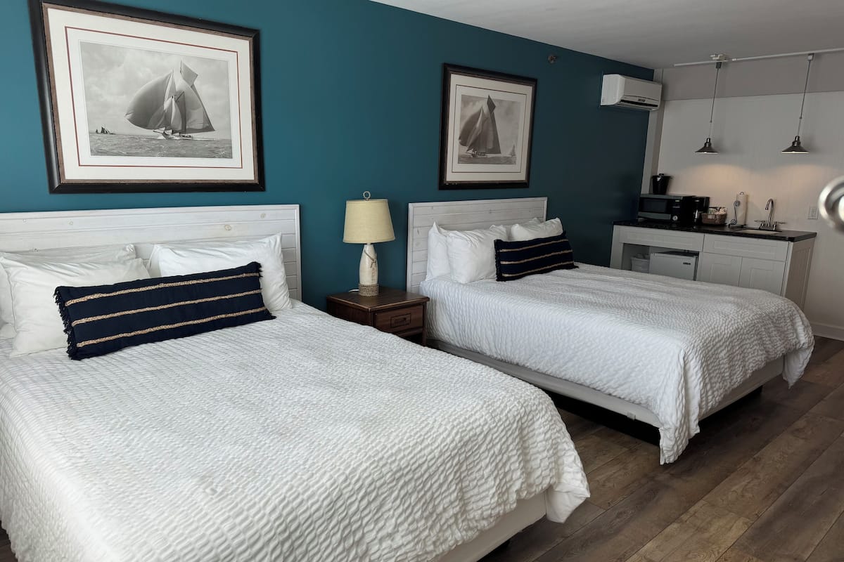 A hotel room with two beds, white bedding, a teal accent wall, framed black and white sailboat pictures, a small bedside table with a lamp, and a kitchenette area.