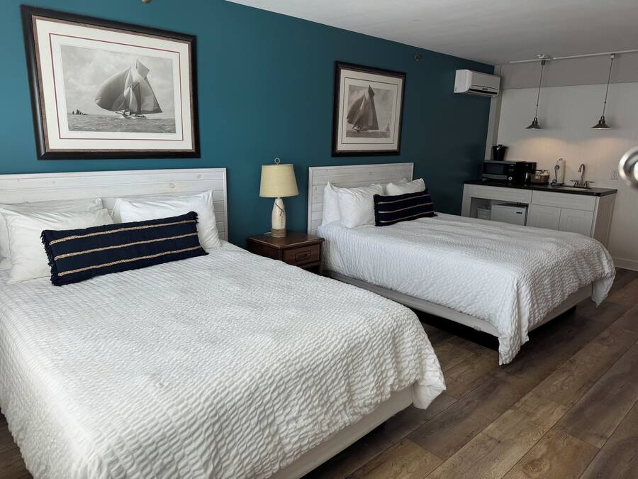 A hotel room with two beds, white bedding, a teal accent wall, framed black and white sailboat pictures, a small bedside table with a lamp, and a kitchenette area.