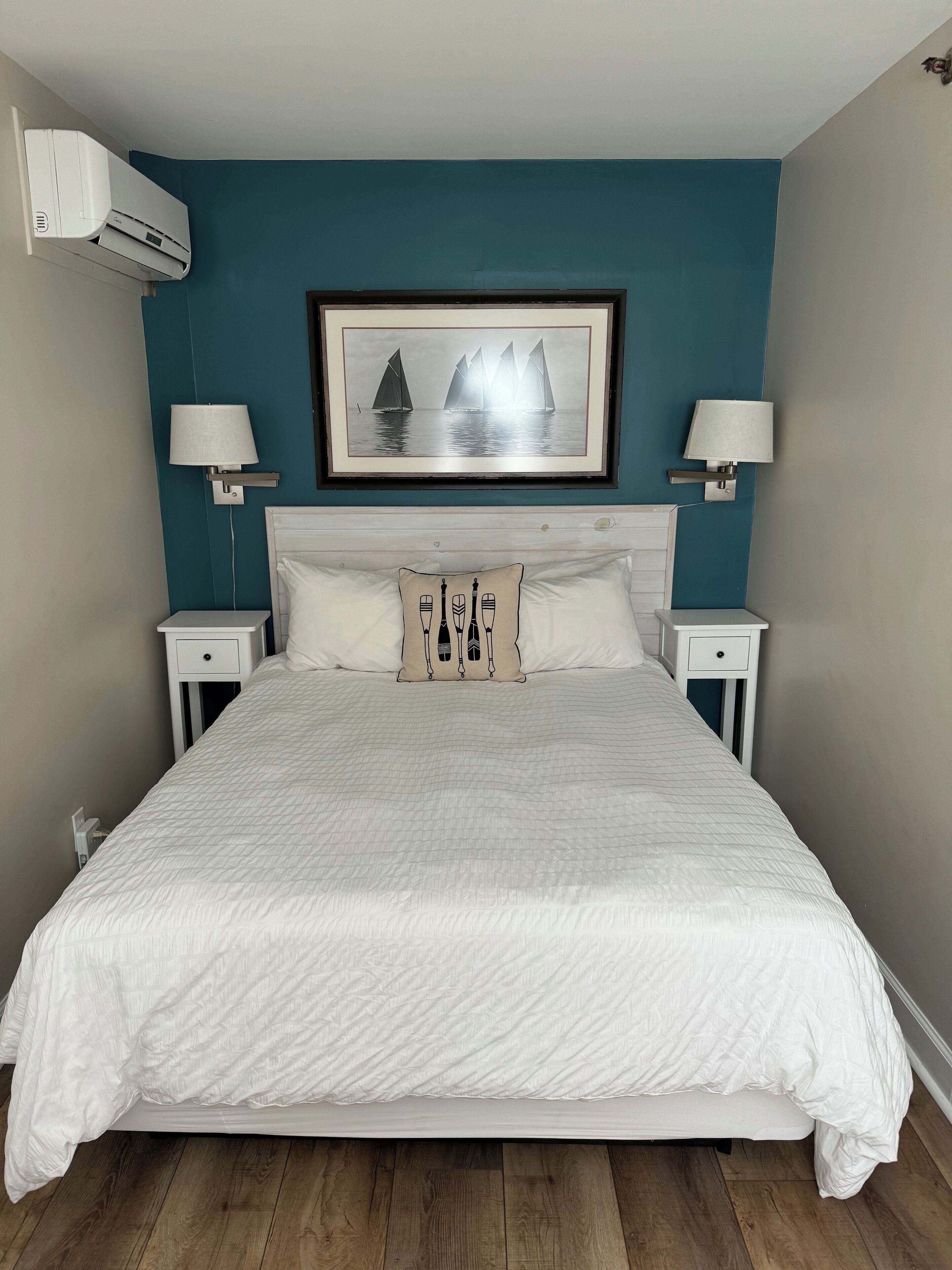 Una cama bien hecha con colcha blanca, dos mesitas de noche con lámparas, un cuadro enmarcado en la pared y un equipo de aire acondicionado en la pared.