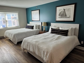 A bedroom with two beds, a window with blinds, a nightstand with a lamp, and framed pictures on the wall. - Pontiac Lodge (Mackinac Island)