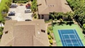 An aerial view of a residential area with houses, a tennis court, and a beach in the background.