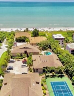 An aerial view of a residential area with houses, a tennis court, and a beach in the background.