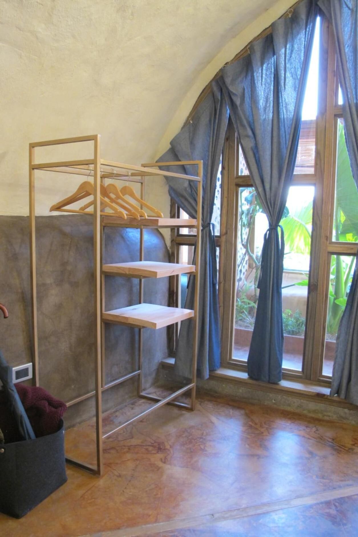 Um quarto com uma estante de madeira encostada à parede, uma janela com cortinas e piso de concreto.
