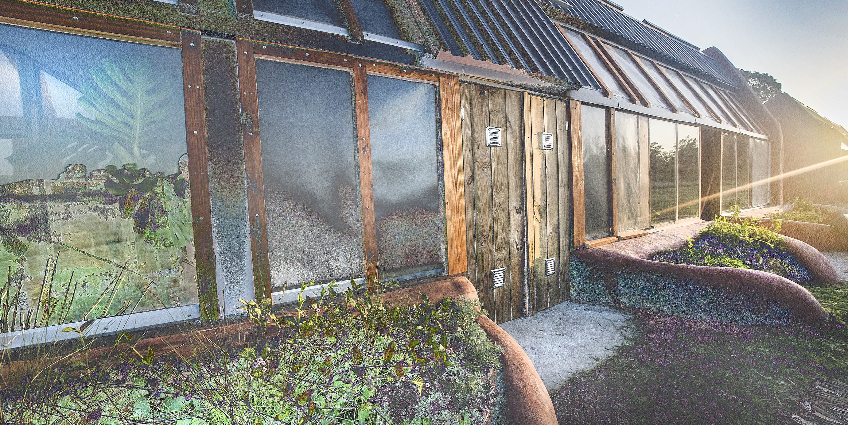 A greenhouse with wooden frames and glass panels, surrounded by plants and a concrete pathway.