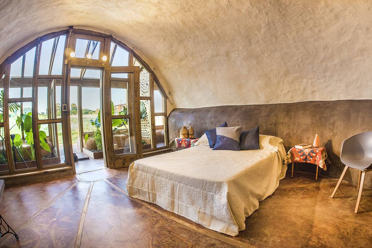 A bedroom with a large arched window, a bed with a patterned bedspread, a bedside table with decorative items, and a modern chair.