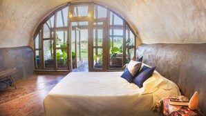 Une chambre confortable avec une grande fenêtre en arc de cercle, un lit recouvert d’une courtepointe blanche et une vue sur la verdure environnante.