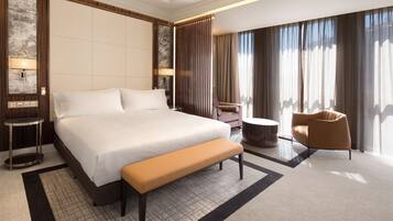 Una habitación de hotel moderna con una cama grande, un banco, una mesa de centro redonda, un sofá y un ventanal con cortinas.