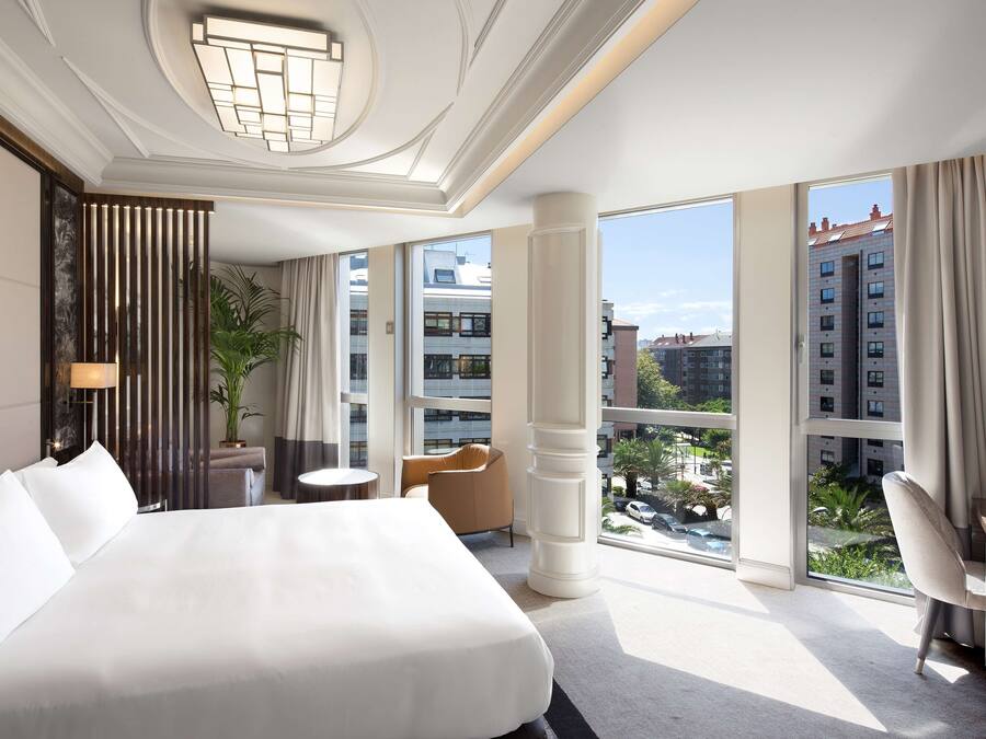 Habitación de hotel con una cama grande, un escritorio, una silla, una planta en maceta y vistas de la ciudad a través de amplios ventanales.