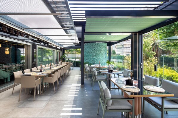 Restaurante moderno com mesas e cadeiras compridas, divisória de vidro e vista para o verde lá fora.