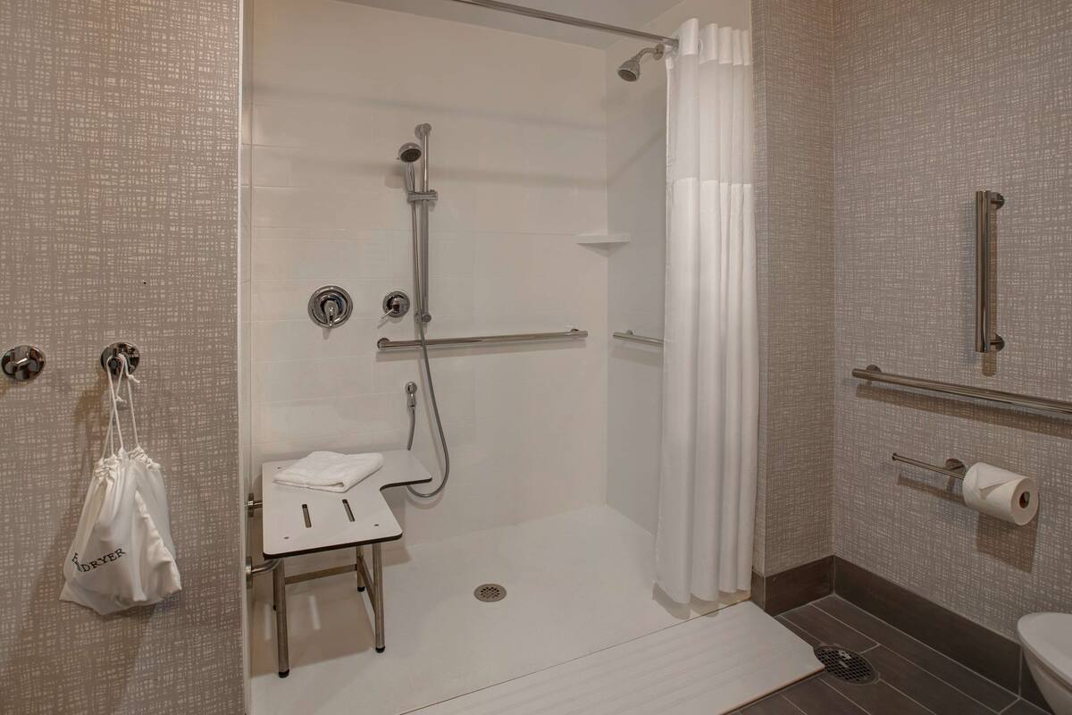 A modern bathroom with a walk-in shower, grab bars, a small table with a towel, and a toilet with a paper towel holder.
