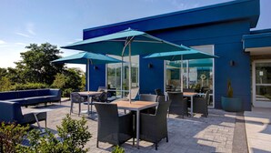 An outdoor patio with blue umbrellas, wicker furniture, and a blue building in the background. - Hampton Inn & Suites Deptford (Woodbury)