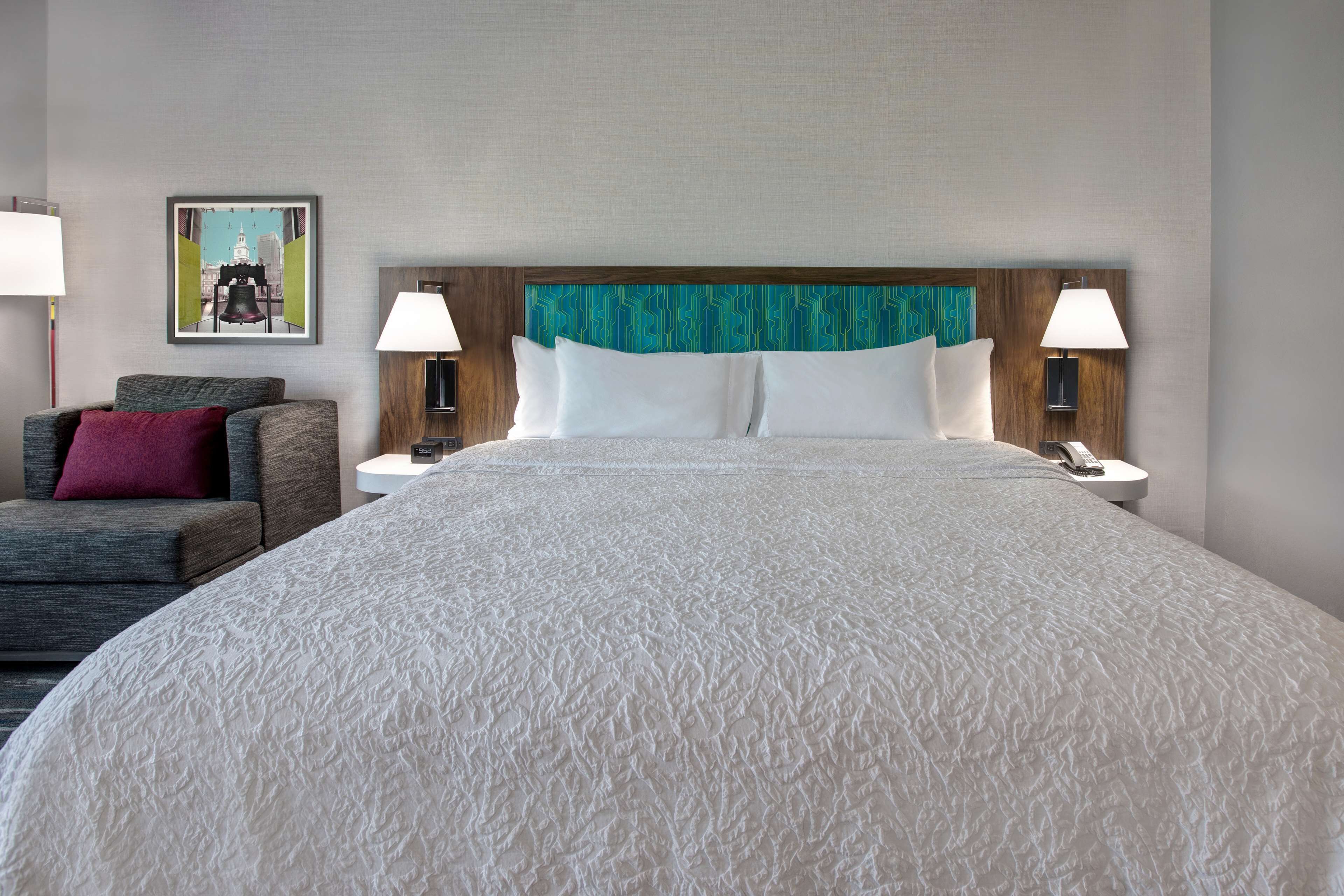 A hotel room with a large bed, two bedside tables with lamps, a gray sofa with a red pillow, and a painting on the wall.