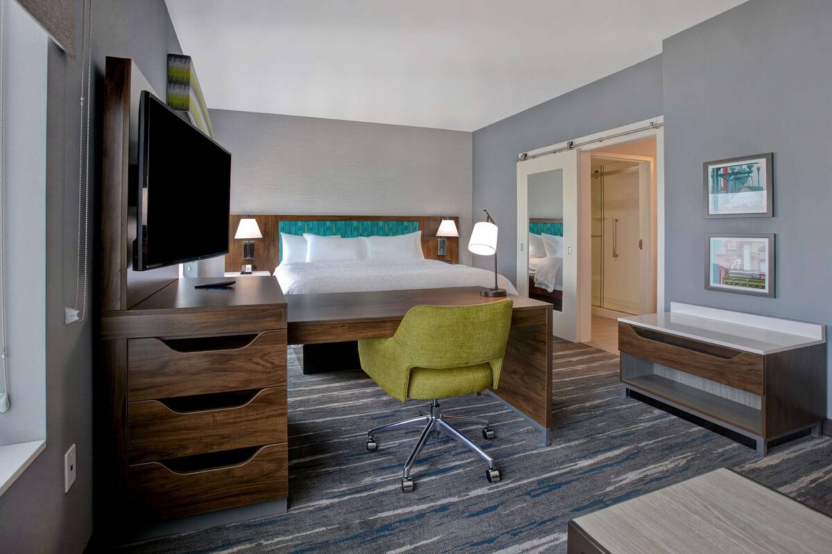 A hotel room with a bed, desk, chair, TV, and artwork on the walls.