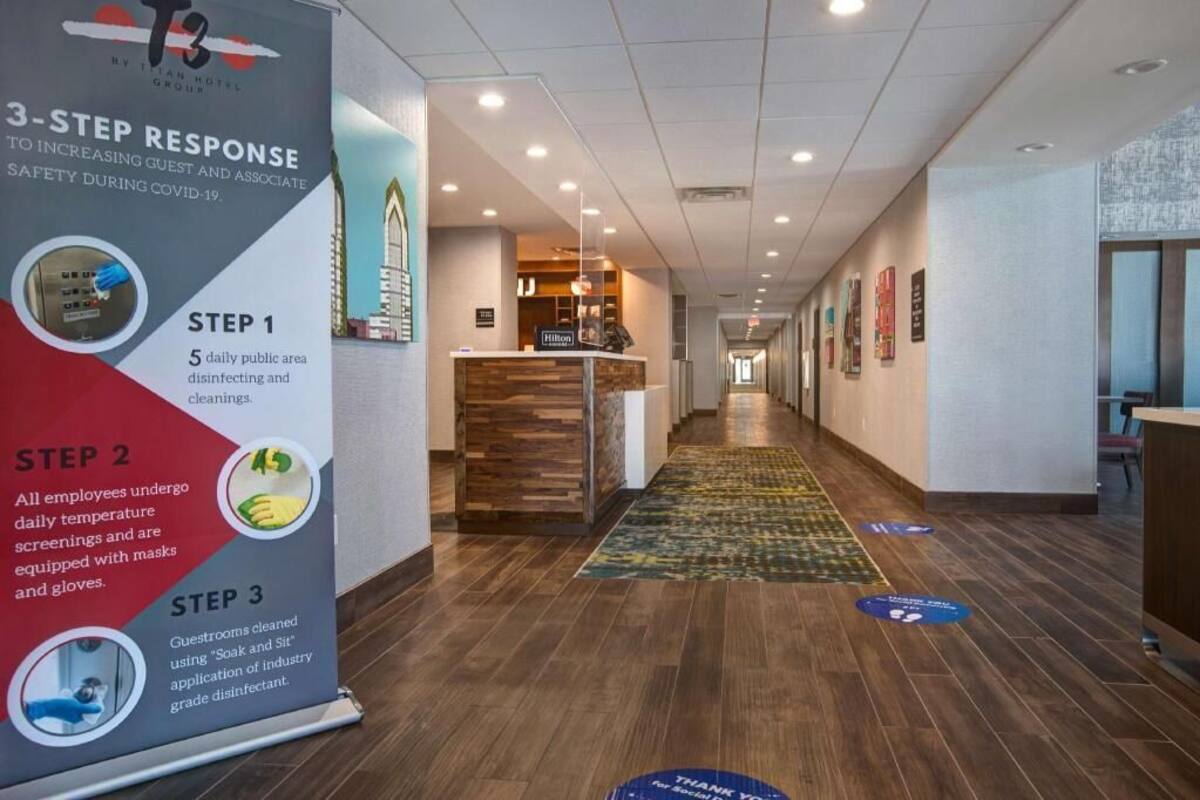 A hotel reception area with a 3-step COVID-19 response poster, a reception desk, and a hallway with a patterned carpet.
