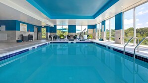 A modern indoor swimming pool with clear blue water, surrounded by blue and white walls, and large windows offering a view of the outdoors. - Hampton Inn & Suites Deptford (Woodbury)