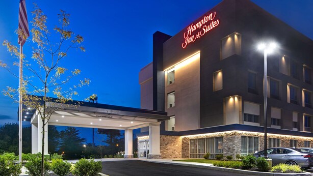 A Hampton Inn & Suites hotel with a clear sky, an American flag, and a well-lit entrance. - Hampton Inn & Suites Deptford (Woodbury)