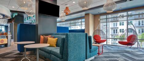Un espace de détente moderne avec un canapé de couleur turquoise, de petites tables, des fauteuils suspendus et une grande fenêtre donnant sur les bâtiments.