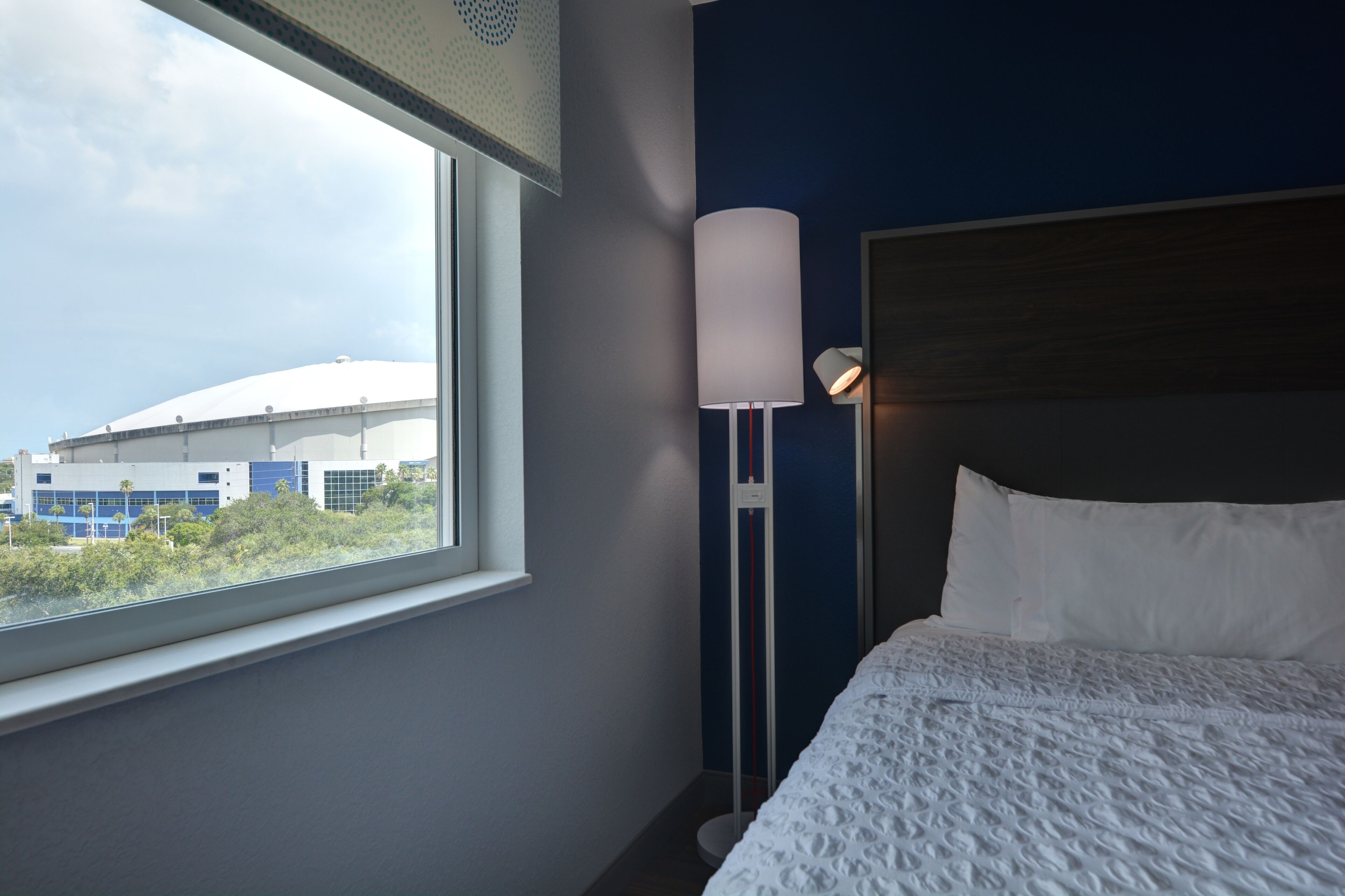 Habitación de hotel con un ventanal grande, cama con ropa de cama blanca, una lámpara de pie alta y vista a un edificio.