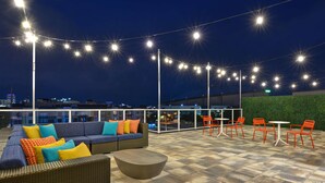 An outdoor seating area with a sofa, chairs, and tables, surrounded by a high wall and string lights. - Tru By Hilton St. Petersburg Downtown Central Ave, FL (St. Petersburg)