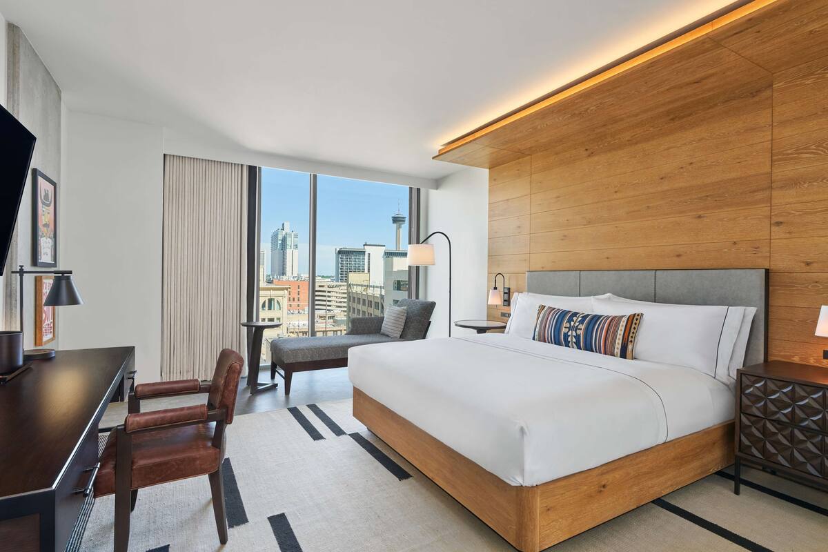 A modern hotel room with a large bed, a desk, and a view of the cityscape.