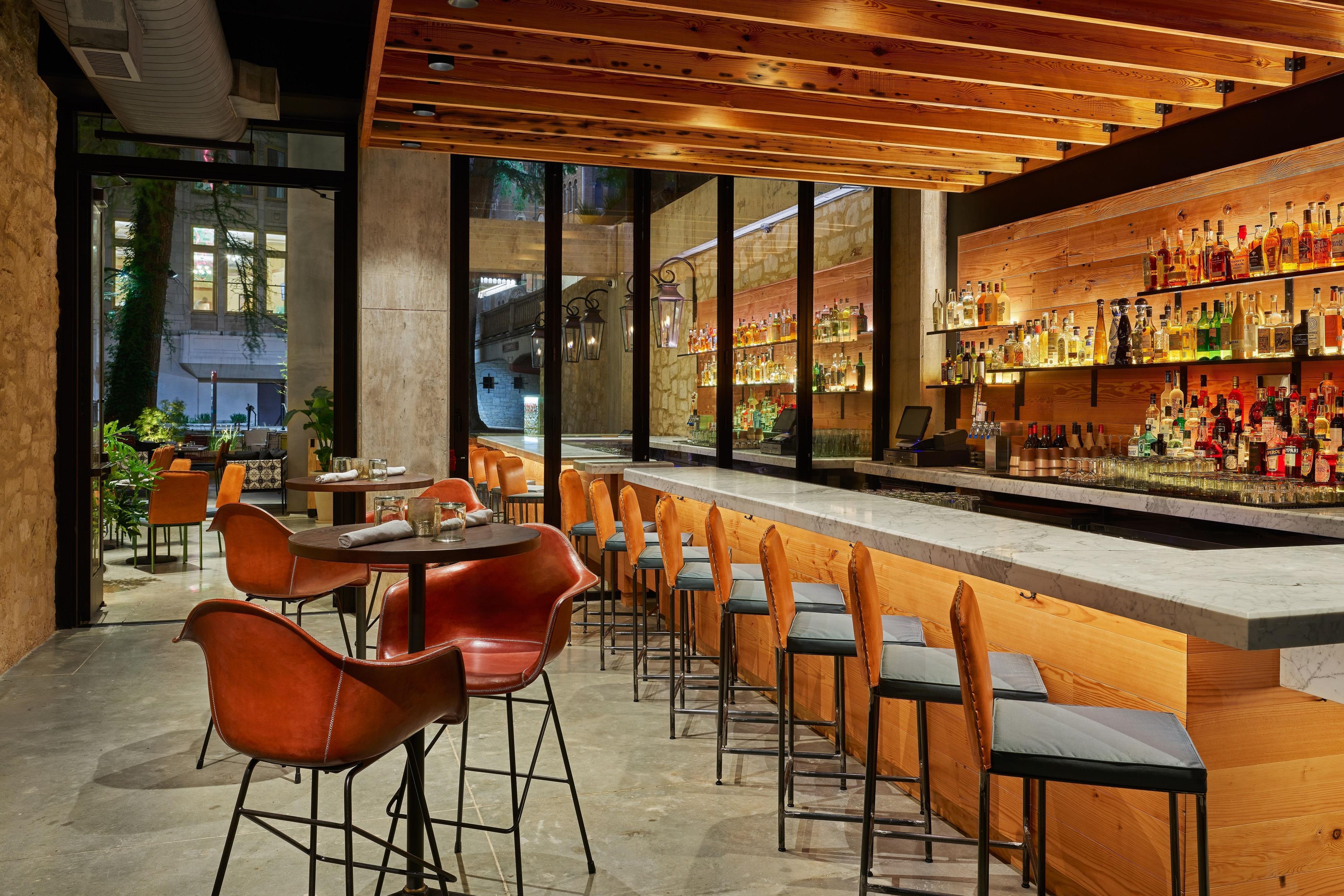 A bar area with wooden shelves stocked with bottles, a seating area with chairs and tables, and large windows offering a view of the outside.