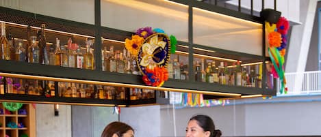 Zona de bar con un bartender y un cliente, decorada con banderines de papel de colores y un ambiente festivo.