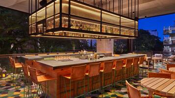 An outdoor bar area with a long counter, multiple bar stools, and a hanging light fixture above.