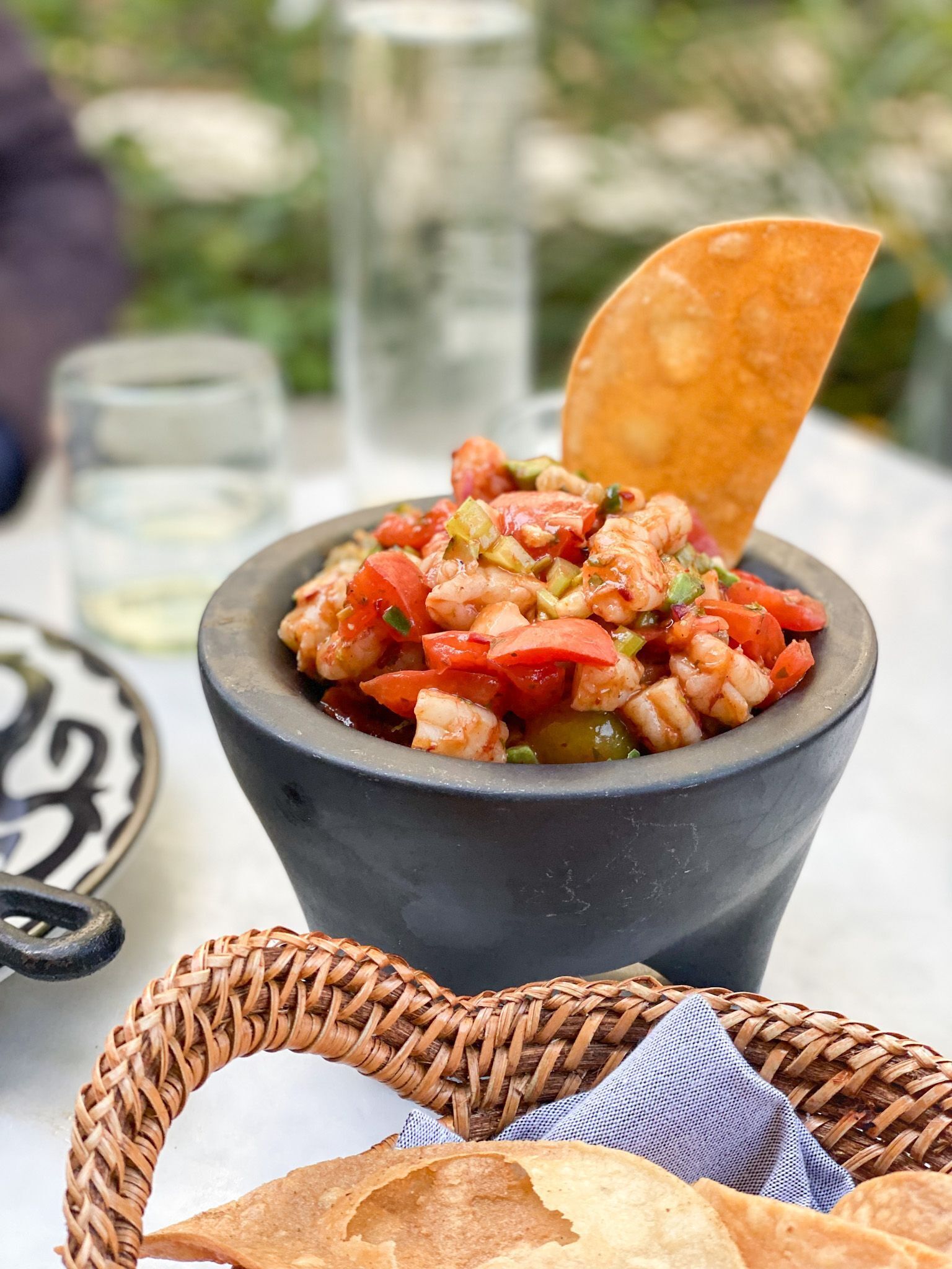A bowl of shrimp and vegetable salad with a tortilla chip on top, placed on a table with a woven basket and a plate nearby.