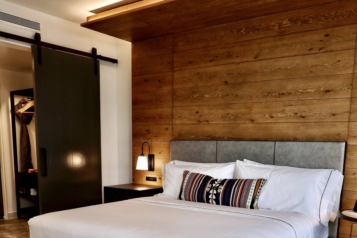 A modern bedroom with a wooden headboard, a large bed with white bedding, a bedside table, and a lamp.