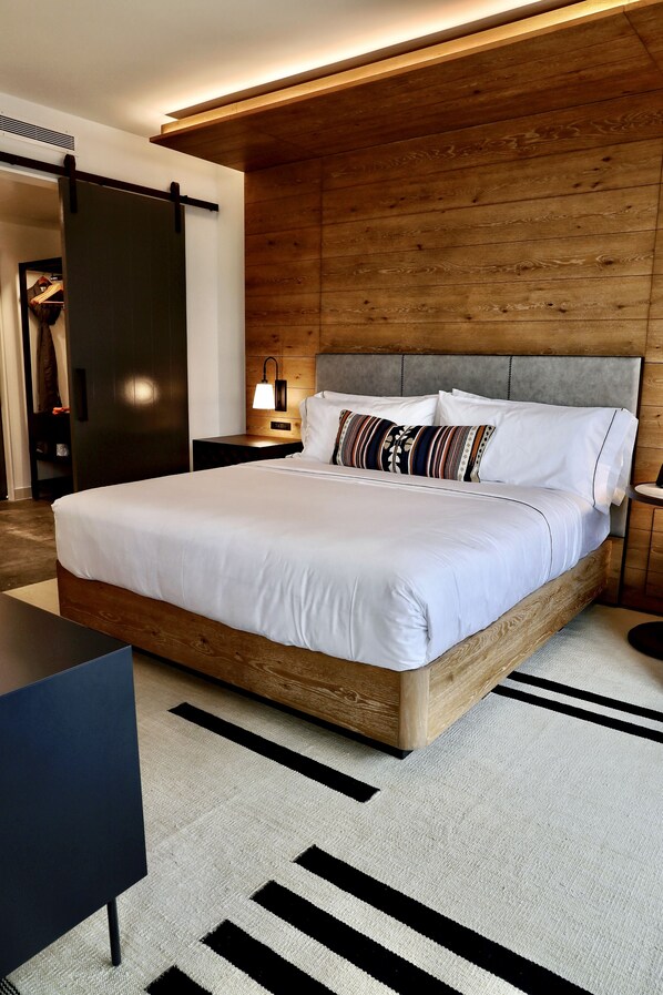 A modern bedroom with a wooden headboard, a large bed with white bedding, a bedside table, and a lamp.