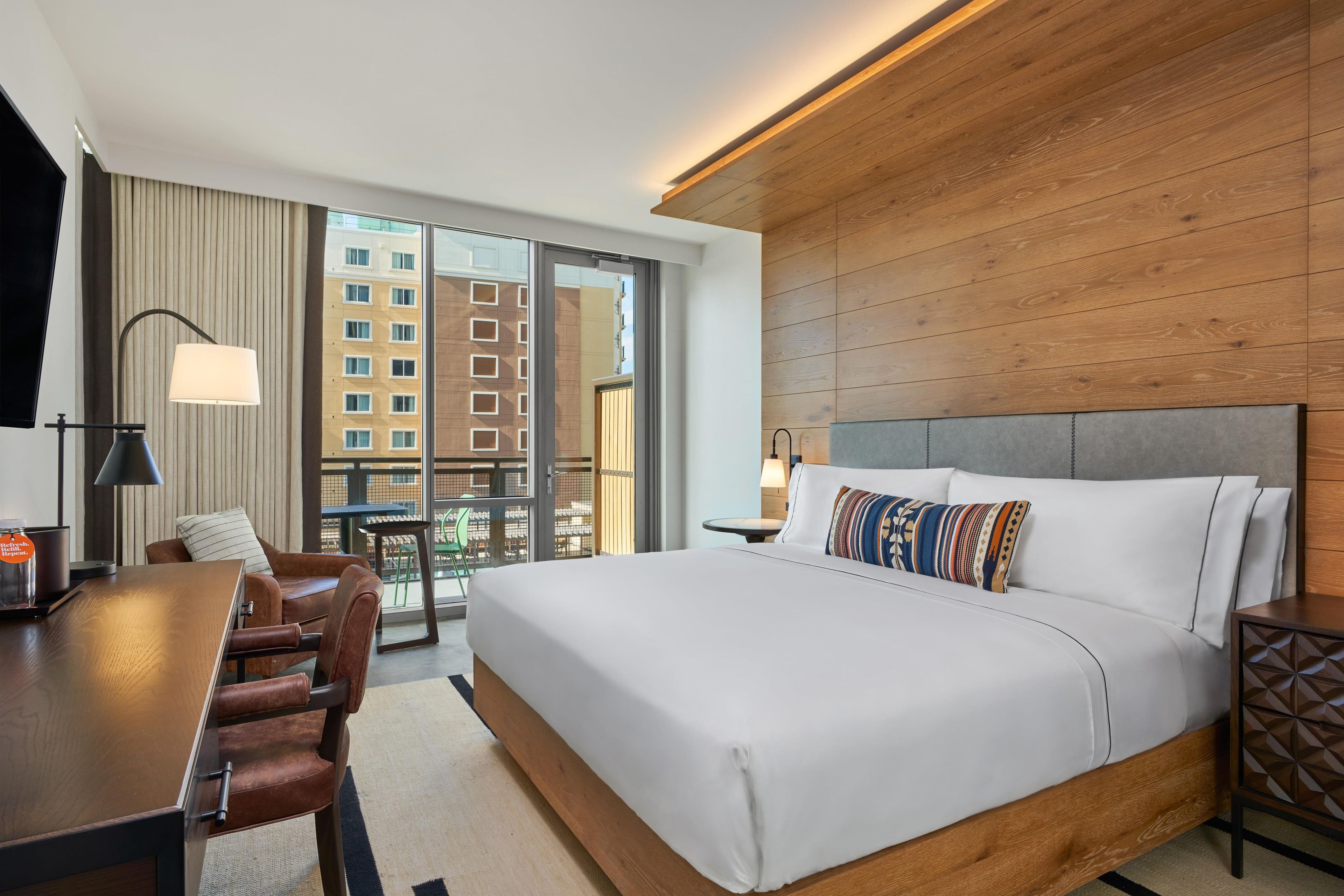Habitación de hotel con una cama grande, un escritorio, una silla, un televisor y vistas a edificios a través de la ventana.