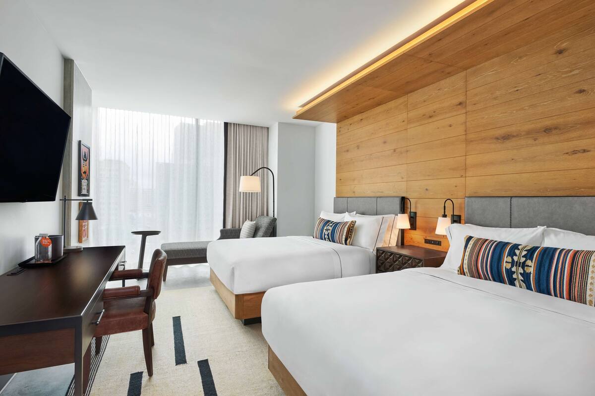 A hotel room with two beds, a desk, a chair, a TV, and a window with curtains.