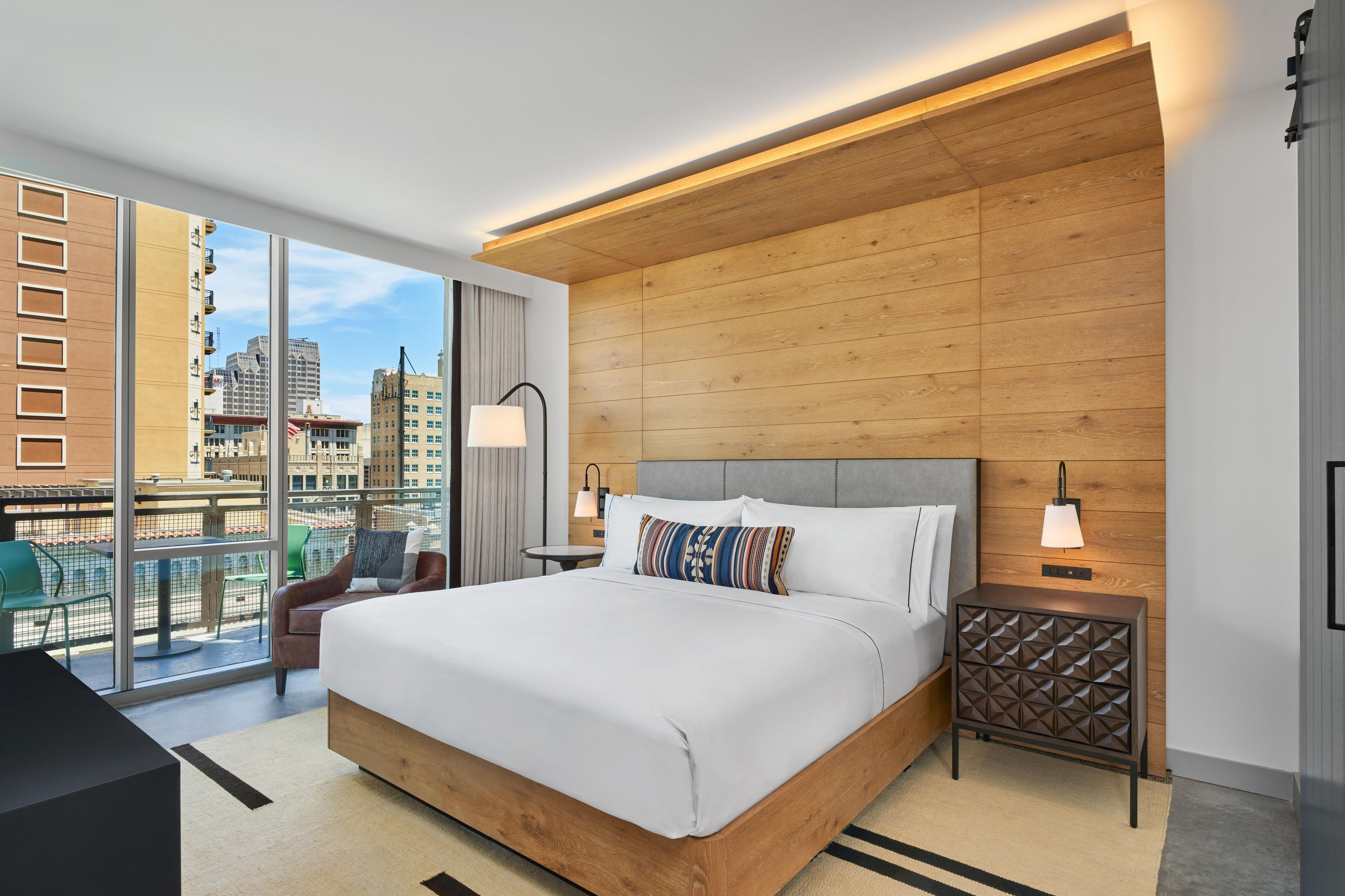 A modern hotel room with a large bed, a view of the city, and a balcony with outdoor seating.