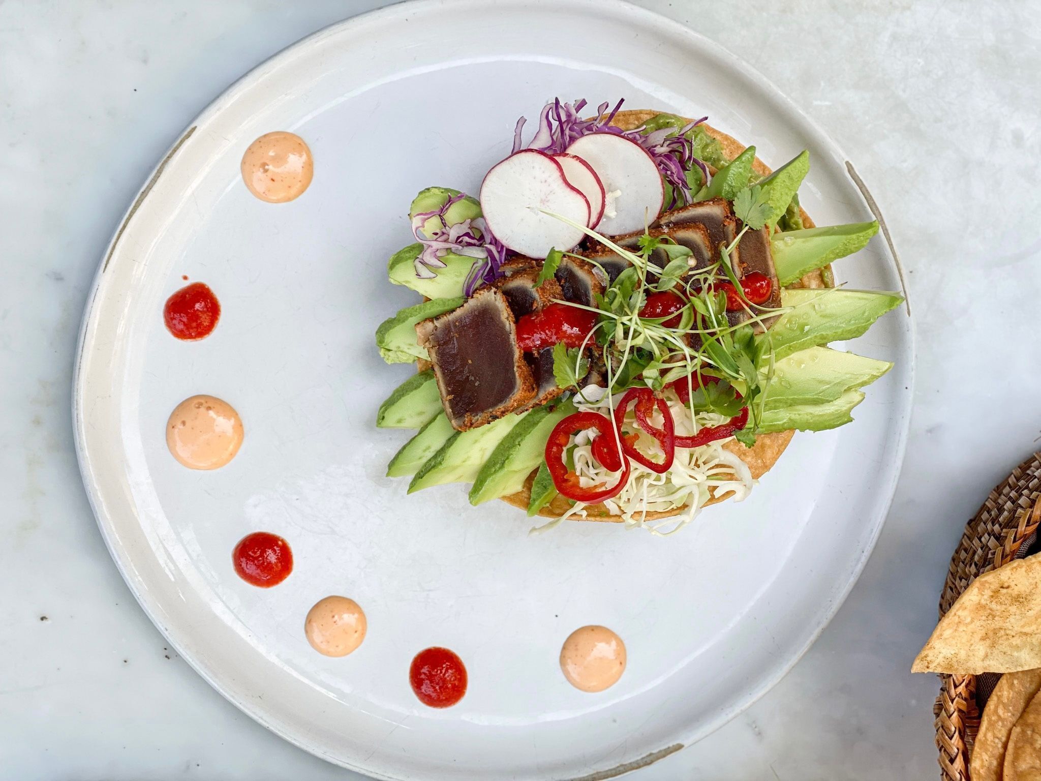 A plate with a variety of food items including avocado, radishes, and sprouts, arranged on a white plate with sauce dots.