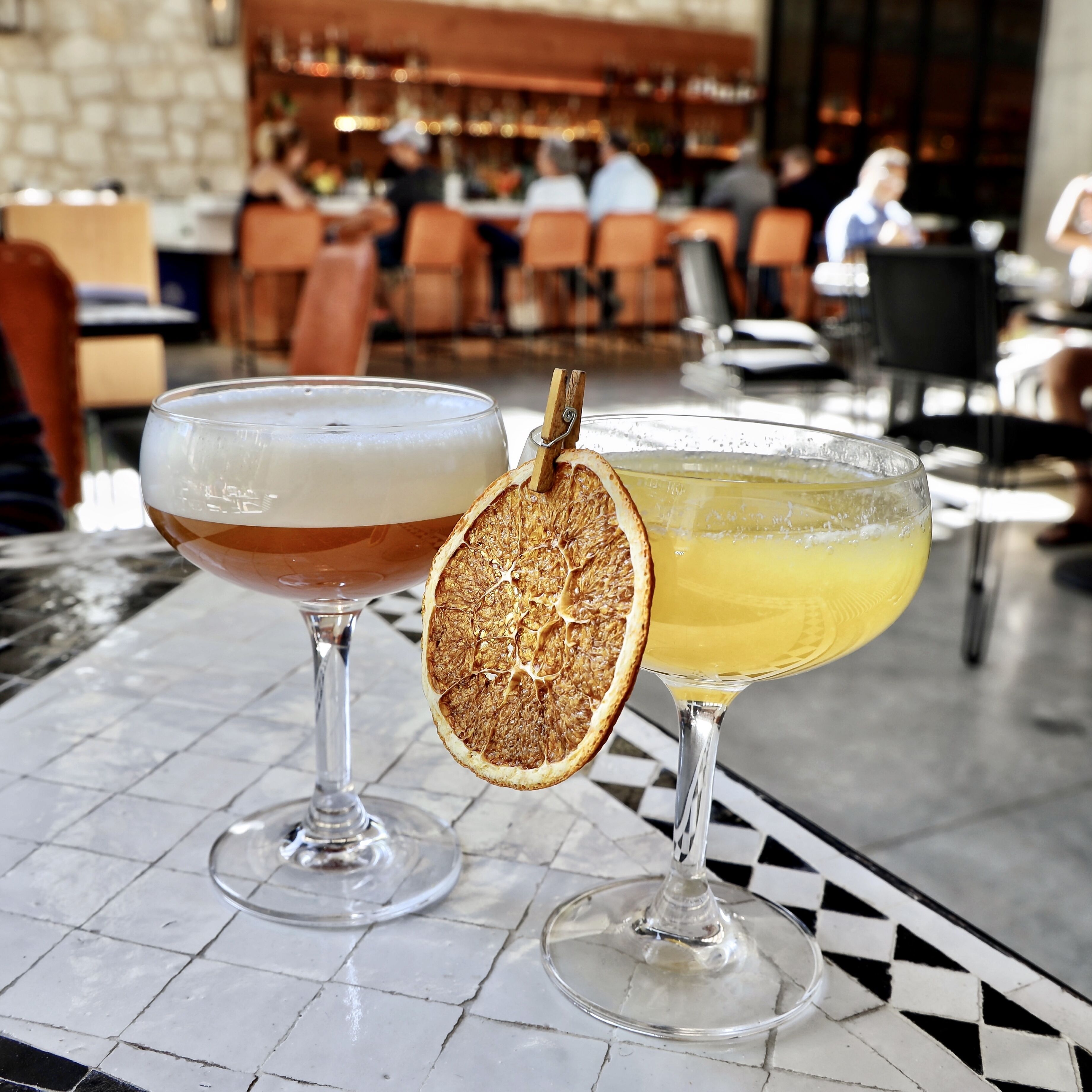 Due cocktail su un tavolo piastrellato, con un bancone bar sullo sfondo.